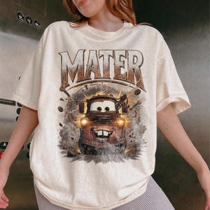 Puede incluir: Camiseta color crema con un gr&aacute;fico de Mate de la pel&iacute;cula Cars. La palabra "MATER" est&aacute; impresa en la parte superior en una fuente marr&oacute;n desgastada. El gr&aacute;fico muestra a Mate con efectos de rayos y humo.