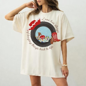 May include: An off-white t-shirt featuring a graphic of a black tire framing a blue sky scene with cartoon characters. The design includes a red cowboy hat, a red cowboy boot labeled Taylor, and the text Love has storys of bringing things back to life.