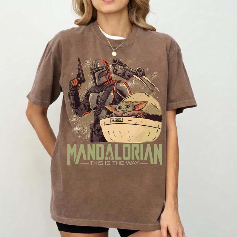 May include: A brown short-sleeve t-shirt featuring a graphic print of a armored character holding a blaster and a small creature in a floating pod. The text MANDALORIAN and THIS IS THE WAY is printed in green below the central character illustration.