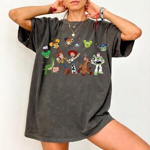 May include: Dark gray t-shirt featuring a colorful graphic of animated characters. The design includes a dinosaur, pig, cowboy, cowgirl, horse, and space ranger. The shirt has a relaxed fit and short sleeves.