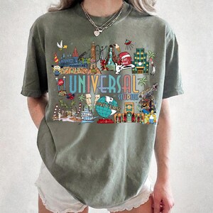 May include: Olive green t-shirt featuring a colorful graphic of Universal Studios attractions. The design includes the words "UNIVERSAL STUDIOS" in a rainbow font, along with various cartoon characters and landmarks. The shirt is a comfortable, casual style.