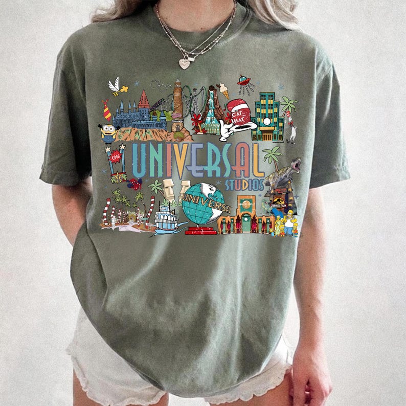 May include: Olive green t-shirt featuring a colorful graphic of Universal Studios attractions. The design includes the words "UNIVERSAL STUDIOS" in a rainbow font, along with various cartoon characters and landmarks. The shirt is a comfortable, casual style.