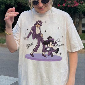 May include: Cream-colored t-shirt featuring a graphic of Michael Jackson and Mickey Mouse in purple suits and hats, dancing on a purple stage. The text "Michael Jackson" and "Mickey Mouse" are also visible.