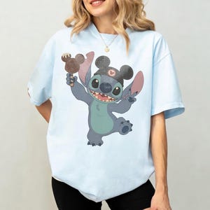 May include: Light blue t-shirt featuring a cartoon Stitch character wearing a Mickey Mouse hat and holding a Mickey Mouse ice cream bar. The character is smiling and giving a peace sign. The shirt is a light blue color.