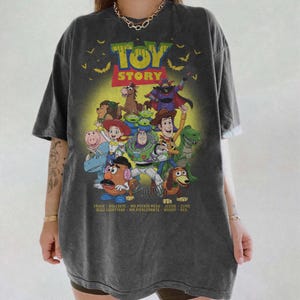May include: A charcoal grey short-sleeve t-shirt featuring a graphic print of Toy Story characters including Buzz Lightyear, Woody, Jessie, and Mr. Potato Head. The design includes the text "TOY STORY" and a list of character names at the bottom.