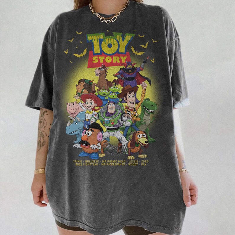 May include: A charcoal grey short-sleeve t-shirt featuring a graphic print of Toy Story characters including Buzz Lightyear, Woody, Jessie, and Mr. Potato Head. The design includes the text "TOY STORY" and a list of character names at the bottom.