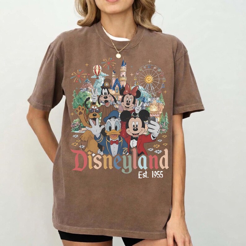 May include: A brown short-sleeve t-shirt featuring a graphic print of Mickey Mouse, Minnie Mouse, Donald Duck, Goofy, and Pluto in front of a Disneyland castle and Ferris wheel. The text reads Disneyland Est. 1955 in a multi-colored vintage font.