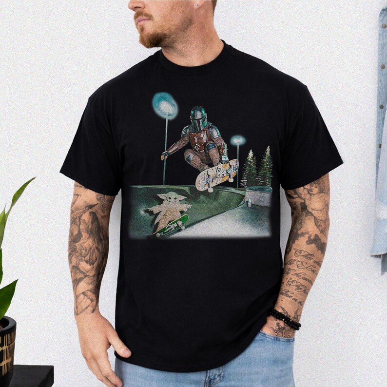 May include: Black t-shirt featuring a graphic of the Mandalorian and Grogu skateboarding in a skate park. The Mandalorian is wearing his signature armor, and Grogu is riding a small skateboard. The background includes trees and streetlights.