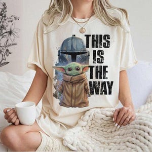 Peut inclure: T-shirt cr&egrave;me avec un motif d'un casque Mandalorien et de Grogu, avec le texte "THIS IS THE WAY" en noir. Le design comprend un casque bleu et un Grogu vert dans un manteau marron.