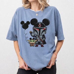 May include: Light blue t-shirt featuring a cartoon illustration of The Mandalorian and Grogu, both with Mickey Mouse ears. The Mandalorian is in his armor, Grogu is in his robe, and a Mickey Mouse balloon floats nearby.