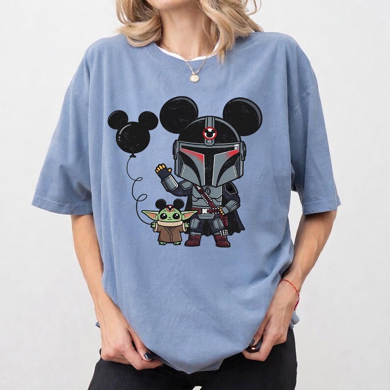May include: Light blue t-shirt featuring a cartoon illustration of The Mandalorian and Grogu, both with Mickey Mouse ears. The Mandalorian is in his armor, Grogu is in his robe, and a Mickey Mouse balloon floats nearby.