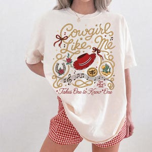 May include: An off-white t-shirt featuring a graphic print with the text Cowgirl Like Me and Takes One to Know One. The design includes a red cowboy hat, a horseshoe with a rose, a cactus, musical notes, and a lasso rope border in yellow.
