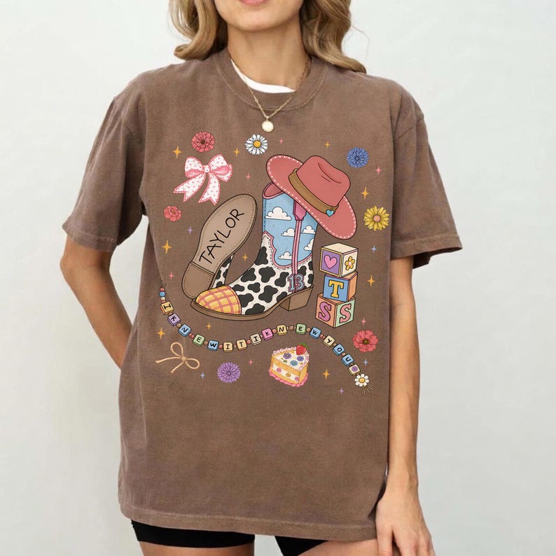 May include: A brown short-sleeve t-shirt featuring a graphic print of a cow-print cowboy boot, a pink hat, alphabet blocks spelling T-S-S, a slice of cake, and a bow. The text TAYLOR is printed on the boot, and the text IN NEW IT K NEW TOUR is on a string.