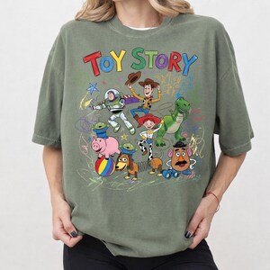 Puede incluir: Camiseta verde oliva con las palabras "TOY STORY" en letras coloridas. La camiseta presenta un gr&aacute;fico de personajes de la pel&iacute;cula animada, incluyendo a Woody, Buzz Lightyear y Rex.