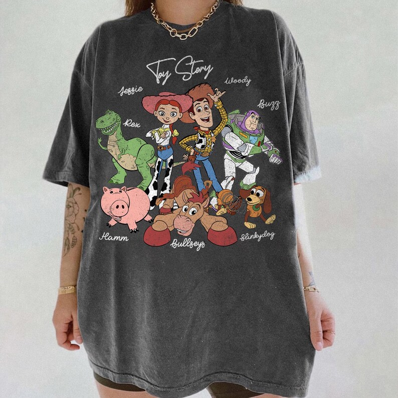 May include: Dark gray t-shirt featuring the words "Toy Story" and cartoon characters Jessie, Rex, Woody, Buzz, Hamm, Bullseye, and Slinky Dog. The shirt has a vintage, worn look.