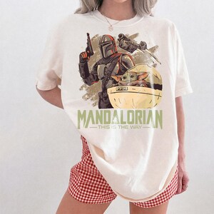 May include: An off-white short-sleeve t-shirt featuring a graphic of a Mandalorian character holding a blaster and a small green creature in a floating pod. The text "MANDALORIAN" and "THIS IS THE WAY" is printed in green below the illustration.