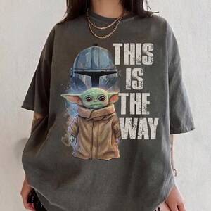 Peut inclure: T-shirt gris fonc&eacute; avec un motif d'un casque et d'un personnage extraterrestre vert, avec le texte "THIS IS THE WAY" en blanc. Le motif est imprim&eacute; sur un t-shirt ample &agrave; manches courtes.