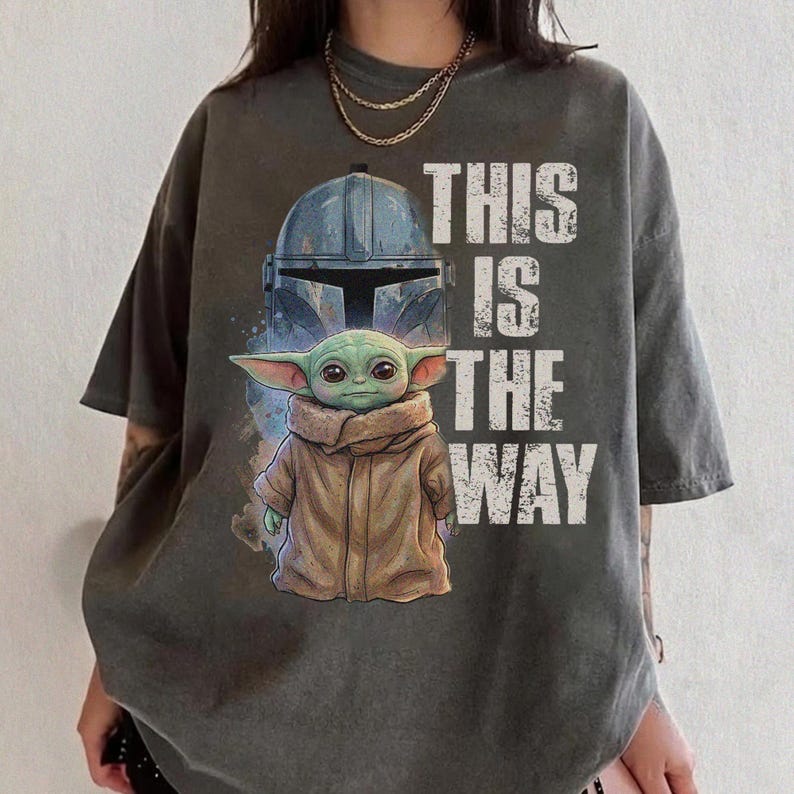 Peut inclure: T-shirt gris fonc&eacute; avec un motif d'un casque et d'un personnage extraterrestre vert, avec le texte "THIS IS THE WAY" en blanc. Le motif est imprim&eacute; sur un t-shirt ample &agrave; manches courtes.
