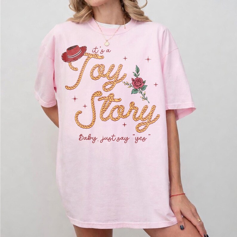 May include: A light pink short-sleeve t-shirt featuring a graphic print with a red cowboy hat, a red rose, and the text "it's a Toy Story" written in a rope-style font, along with the additional text "Baby, just say "yes"" in red script.