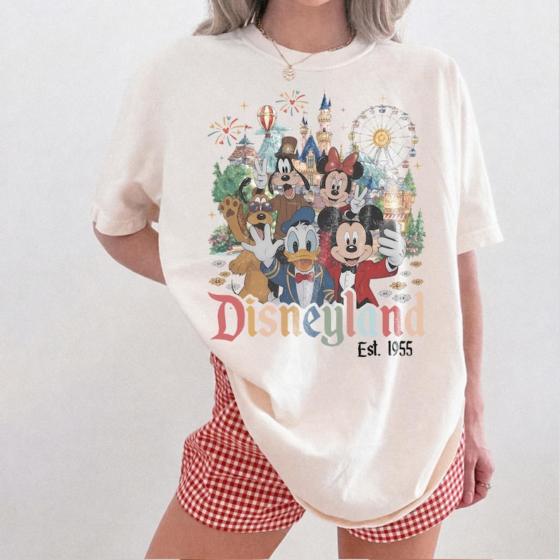 May include: An off-white short-sleeve t-shirt featuring a colorful graphic of Mickey Mouse, Minnie Mouse, Donald Duck, Goofy, and Pluto in front of a castle and Ferris wheel. The text reads Disneyland Est. 1955 in a multi-colored, vintage-style font.