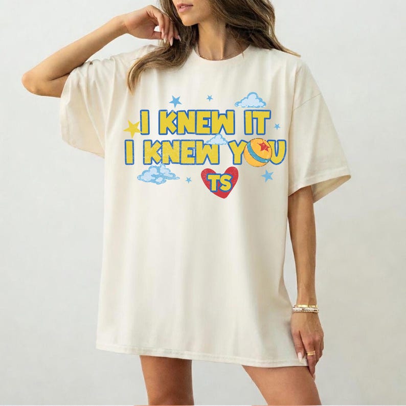 May include: An off-white short-sleeve t-shirt featuring the blue and yellow text I KNEW IT I KNEW YOU TS. The design includes a red heart, a yellow and blue ball, light blue clouds, and small blue stars printed on the front of the garment.