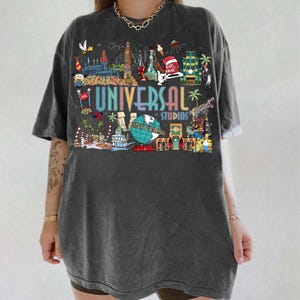 May include: Dark gray t-shirt featuring a colorful graphic of Universal Studios attractions. The design includes the text "UNIVERSAL STUDIOS" in a rainbow font, along with various illustrations of theme park elements.