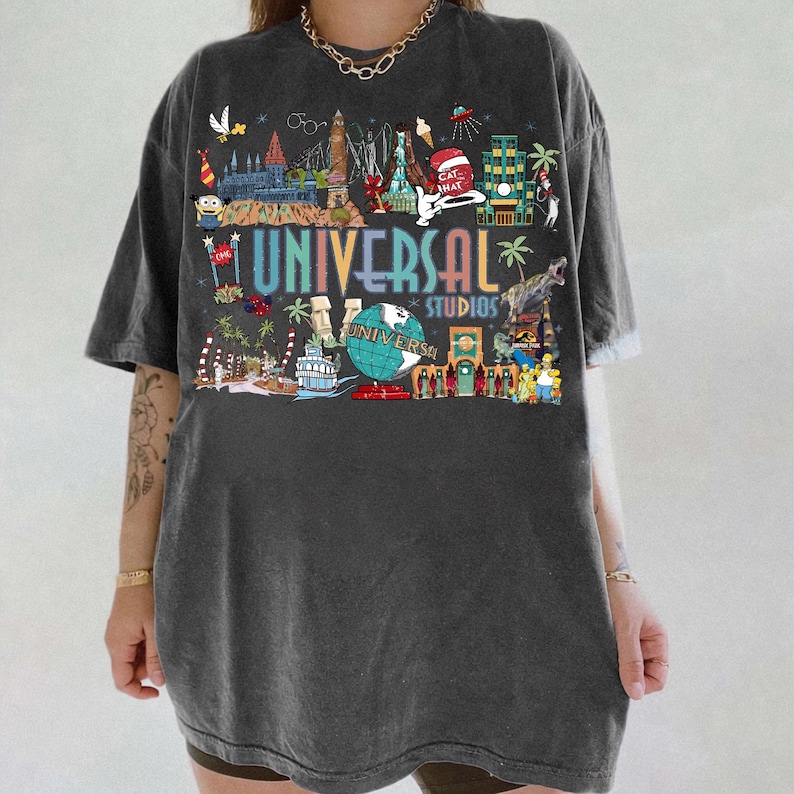 May include: Dark gray t-shirt featuring a colorful graphic of Universal Studios attractions. The design includes the text "UNIVERSAL STUDIOS" in a rainbow font, along with various illustrations of theme park elements.