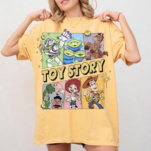 May include: Yellow t-shirt featuring a colorful graphic of characters from the animated movie "Toy Story." The design includes Buzz Lightyear, the aliens, Woody, and other characters. The words "TOY STORY" are printed in large, yellow letters.