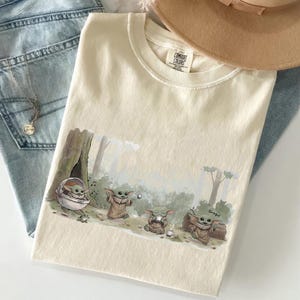 May include: Cream-colored t-shirt featuring a Grogu graphic in a forest scene. The graphic shows Grogu in various poses, including one in a pod and another holding a cup. The shirt is made of Comfort Colors material.