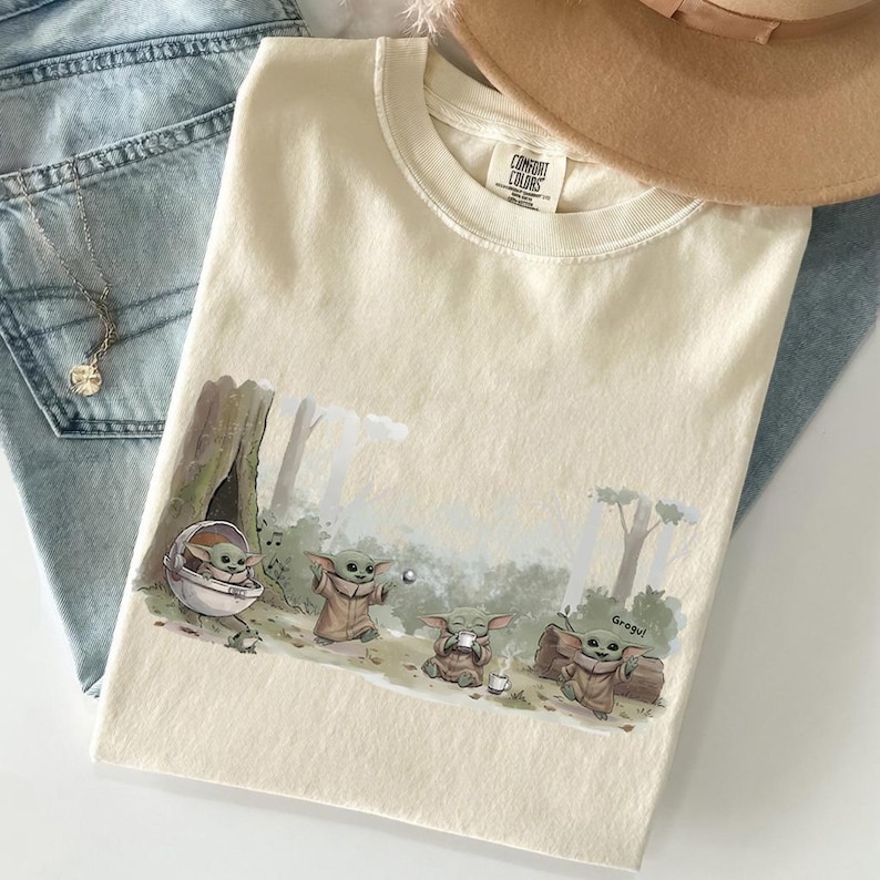 May include: Cream-colored t-shirt featuring a Grogu graphic in a forest scene. The graphic shows Grogu in various poses, including one in a pod and another holding a cup. The shirt is made of Comfort Colors material.