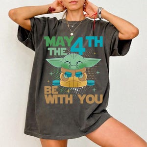 K&ouml;nnte beinhalten: Dunkelgraues T-Shirt mit einer Grogu-Grafik und dem Text "MAY THE 4TH BE WITH YOU" in Gr&uuml;n, Blau und Braun. Das Design zeigt ein Cartoon-Bild von Grogu, auch bekannt als Baby Yoda, der eine Tasse h&auml;lt.