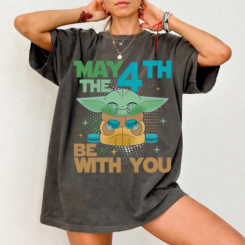 K&ouml;nnte beinhalten: Dunkelgraues T-Shirt mit einer Grogu-Grafik und dem Text "MAY THE 4TH BE WITH YOU" in Gr&uuml;n, Blau und Braun. Das Design zeigt ein Cartoon-Bild von Grogu, auch bekannt als Baby Yoda, der eine Tasse h&auml;lt.