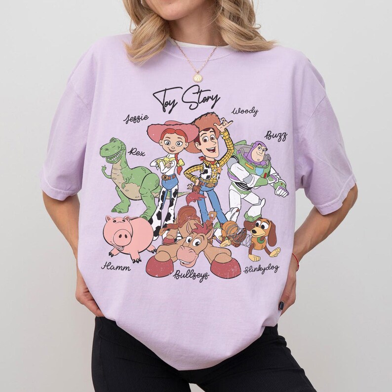 May include: A light purple t-shirt featuring the words "Toy Story" above a graphic of the characters Woody, Buzz Lightyear, Jessie, Rex, Hamm, Bullseye, and Slinky Dog. The shirt is a crew neck and has short sleeves.
