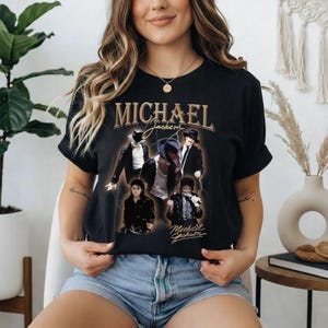 May include: Black t-shirt featuring a collage of images of Michael Jackson, with the name "MICHAEL" in gold lettering at the top. The design includes various poses of the singer, with his signature below.