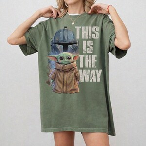 Peut inclure: T-shirt vert olive avec un motif repr&eacute;sentant un casque Mandalorien et Grogu, avec le texte "THIS IS THE WAY" en blanc. Le design comprend une illustration d&eacute;taill&eacute;e de Grogu dans un manteau marron. Le t-shirt est de coupe ample.
