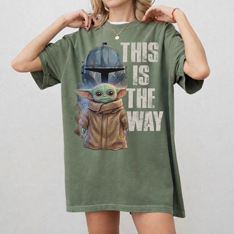 Peut inclure: T-shirt vert olive avec un motif repr&eacute;sentant un casque Mandalorien et Grogu, avec le texte "THIS IS THE WAY" en blanc. Le design comprend une illustration d&eacute;taill&eacute;e de Grogu dans un manteau marron. Le t-shirt est de coupe ample.