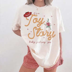 May include: An off-white t-shirt featuring a red cowboy hat, a pink rose, and the text "it's a Toy Story" in a rope-style font, with the additional text "Baby, just say "yes"" printed in red below the main graphic design.