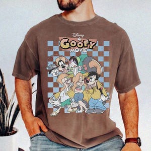 May include: Brown t-shirt featuring a graphic from Disney's "A Goofy Movie." The design includes Goofy, Max, and other characters against a checkered background. The shirt has short sleeves and a crew neck.