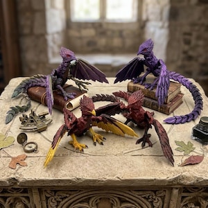 May include: A collection of fantasy creature figurines in shades of purple, red, and yellow. The creatures have wings and are posed on books and a stone surface. Additional props include a compass, inkwell, and decorative leaves.