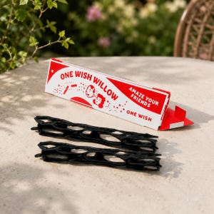 May include: Two black, elongated, organic-shaped plastic structures with hollow cutouts, accompanied by a white and red rectangular cardboard box labeled "ONE WISH WILLOW" and "AMAZE YOUR FRIENDS" with illustrations of two faces and stars.