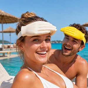 May include: A visor shaped like a human hand, with one version in matte off-white and another in bright yellow. The visor features a molded hand design that rests across the forehead, secured by a matching strap that wraps around the head.