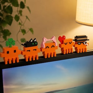 May include: Five orange pixel-art figures lined up on a monitor edge. The first has a green clover, the second a black cat, the third pink cat ears, the fourth a red heart, and the fifth a black hat with sunglasses and a magnifying glass.