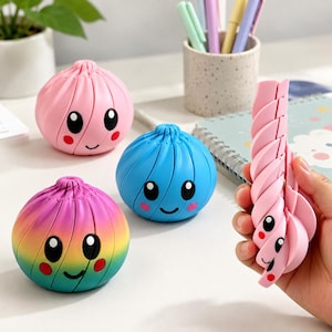 Op de afbeelding: Drie ronde, gesegmenteerde fidget-speeltjes in de vorm van dumplings met lachende gezichtjes. Eén is effen roze, één is effen blauw en één heeft een regenboogverloop. Een vierde roze, gesegmenteerd speeltje wordt vastgehouden, wat de flexibele structuur toont.