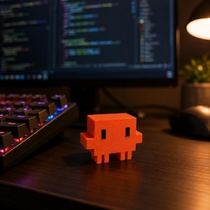 May include: A small, bright orange, three-dimensional pixelated character figurine with a blocky, rectangular head and body, featuring two simple black square eyes and two small protruding arms, standing on two short, blocky legs.
