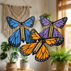 May include: Three butterfly-shaped toys with black frames and translucent wings in blue, purple, and orange. Each toy features a central mechanism with a visible yellow rubber band connecting the upper and lower wing sections for movement.