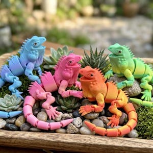 Peut inclure: Quatre figurines d'iguanes articulés aux couleurs bleu, rose, orange et vert sont disposées sur un lit de petits cailloux et de verdure. Les iguanes ont la gueule ouverte et des corps segmentés, suggérant qu'il s'agit de jouets ou d'objets décoratifs.