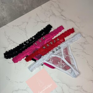 Op de afbeelding: Een verzameling gepersonaliseerde string-slips in zwart, roze, rood en wit. Elke string heeft een naam die met glinsterende strass-steentjes is gespeld. De witte string heeft een bloemenontwerp. Het logo van de lingeriewinkel staat in de hoek.