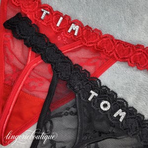 Può includere: Lingerie rossa e nera con finiture in pizzo. La lingerie rossa ha il nome "TIM" scritto in lettere argentate. La lingerie nera ha il nome "TOM" scritto in lettere argentate. La lingerie è su una superficie grigia.