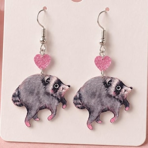 May include: A pair of dangle earrings featuring a gray raccoon illustration with pink accents on the paws and ears. Each earring includes a small, glittery pink heart charm suspended above the raccoon, attached to a silver-toned metal hook.