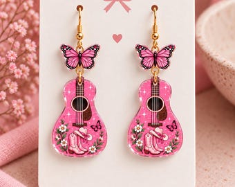 May include: A pair of dangle earrings featuring a pink butterfly charm attached to a pink acoustic guitar charm. The guitar design includes white flowers, a pair of pink cowboy boots, and a small butterfly. The earrings hang from gold-toned hooks.
